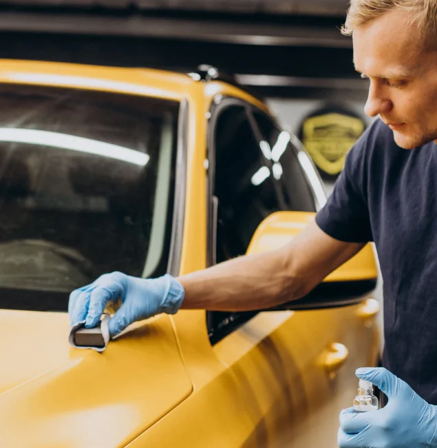 Man wearing blue gloves applying ceramic coating to the hood of a yellow car using an applicator pad.