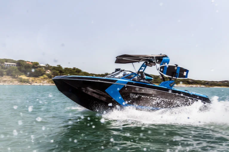 A sleek black and blue wakeboard boat speeding across a lake, creating a splash with water droplets in the foreground and a shoreline in the background.