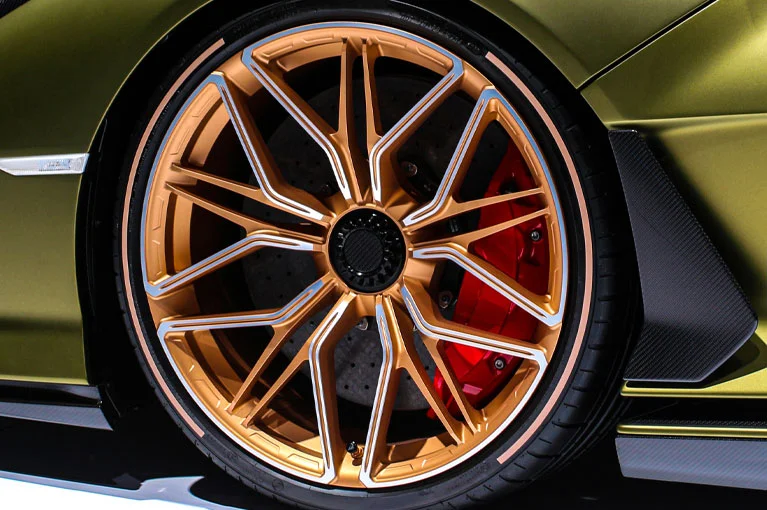 Close-up of a gold alloy wheel with a multi-spoke design on a high-performance sports car, featuring a red brake caliper and carbon fiber detailing around the wheel arch.