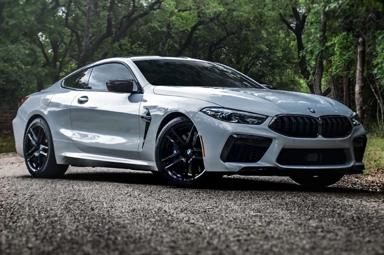 Silver BMW coupe with black alloy wheels parked on a shaded road surrounded by trees.