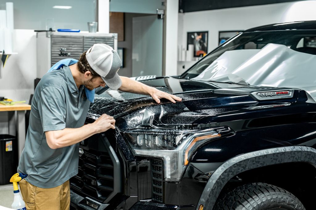 A technician carefully applies paint protection film to the hood and headlight area of a black Toyota Tundra TRD Pro truck. The transparent film is being smoothed out to eliminate bubbles and ensure a seamless fit.