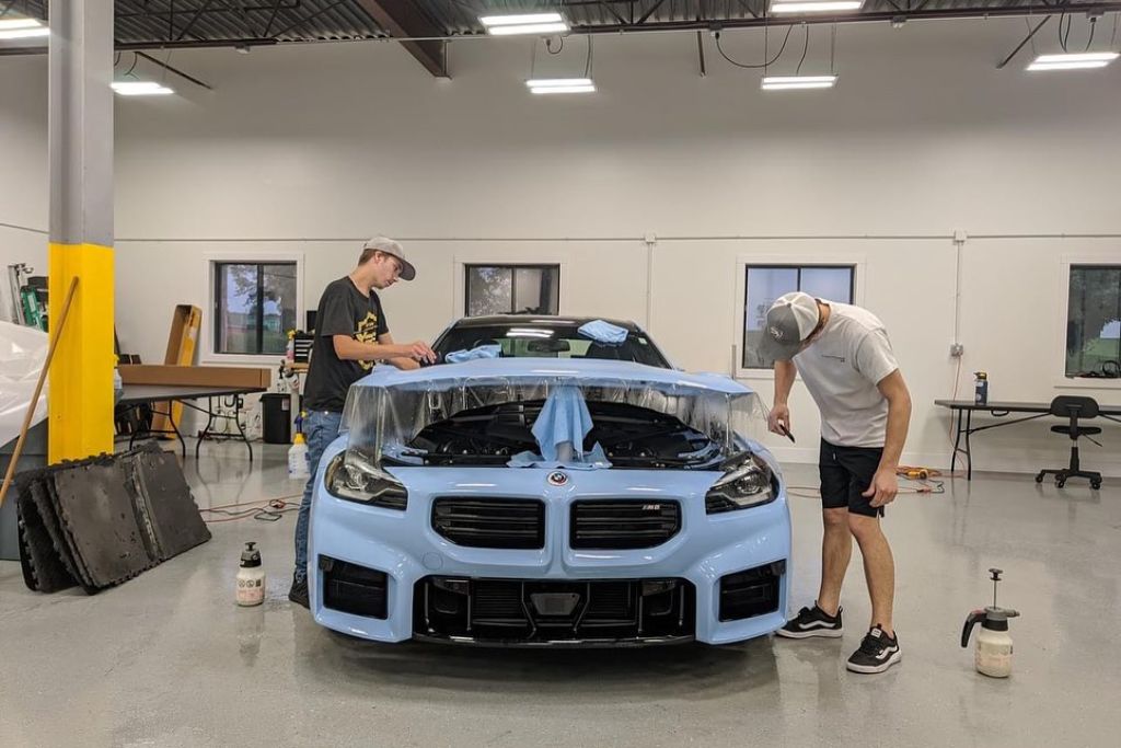Two technicians apply paint protection film to the front of a light blue sports car in a professional garage. The transparent film is draped over the hood and headlights as they work carefully to smooth and secure it.