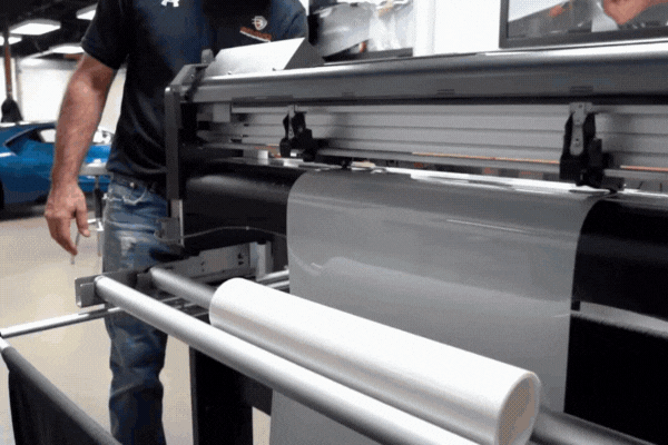 A technician operates a cutting machine that feeds a roll of transparent paint protection film, preparing it for application on a vehicle. The clear film is unrolling smoothly, showcasing its flexibility and precision cutting process.