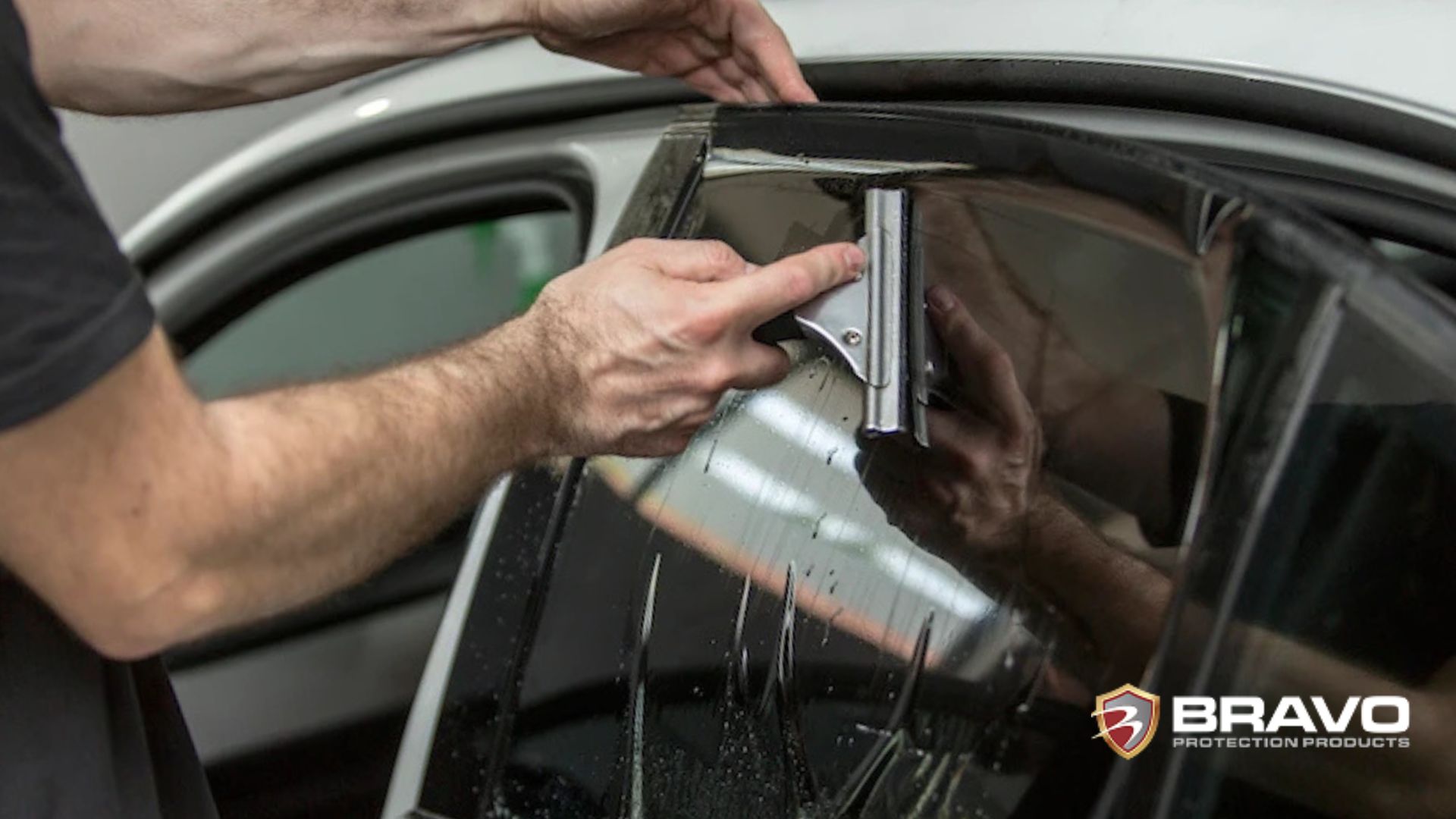 A technician presses a squeegee against a wet window tint film on a car door while aligning and adhering the film into place.