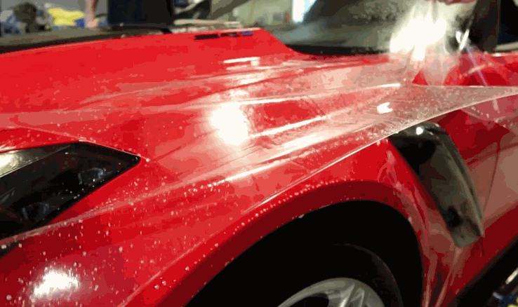 Clear paint protection film being applied to the front fender of a red sports car during PPF installation.