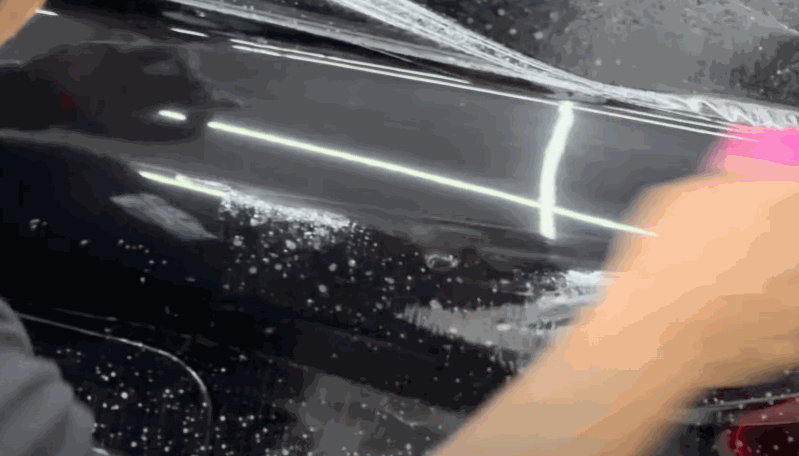A close-up of a worker smoothing paint protection film on a wet black car surface using a pink squeegee.