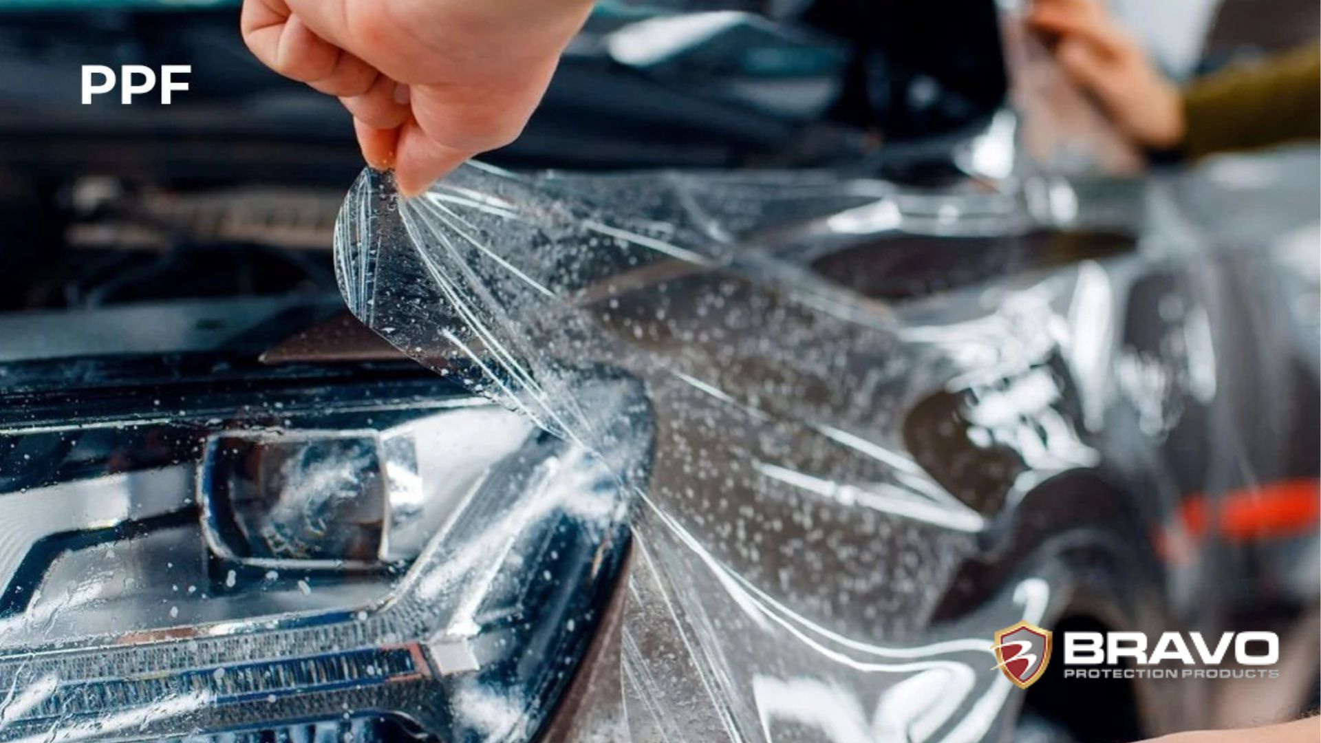 A close-up of a hand lifting a corner of freshly applied paint protection film from a car headlight, with visible water droplets underneath.