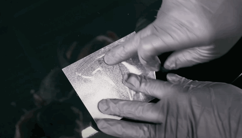 A gloved technician presses down on a small square of shimmering protective film to demonstrate its texture and adhesion.