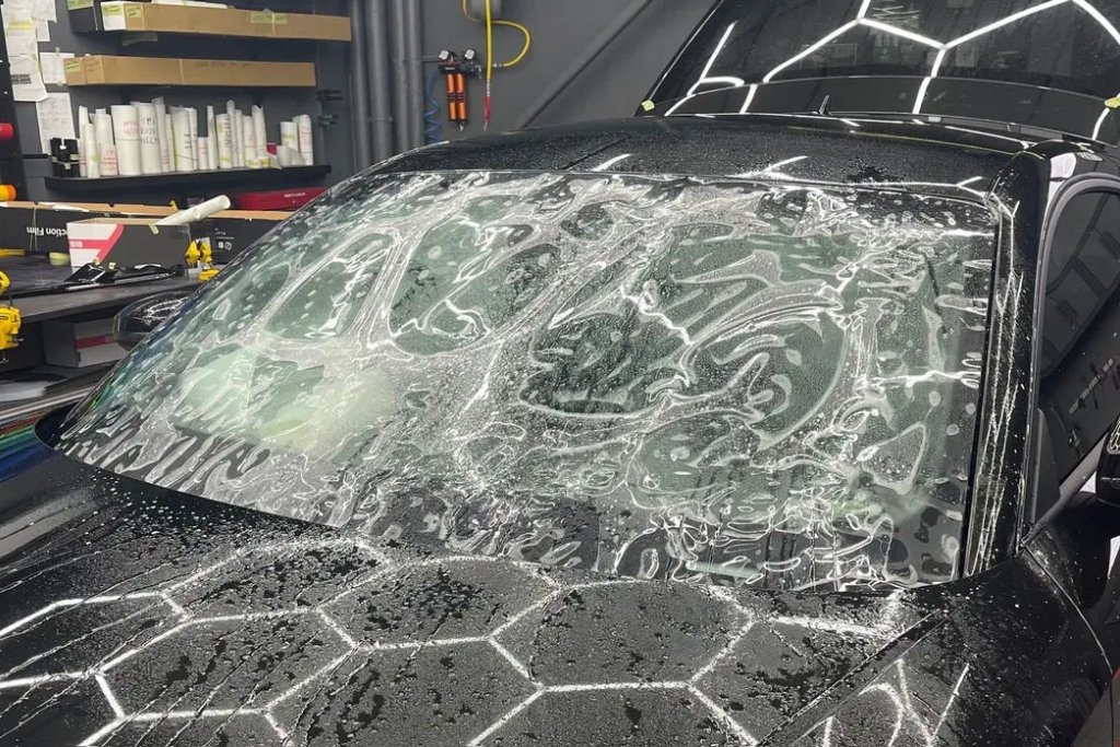 A freshly installed windshield protection film is visible on a black car, covered in application solution and air bubbles as it sets in a detailing shop. This in-progress installation illustrates the maintenance and durability concerns addressed in the guide on How Long Does Windshield Protection Film Last.