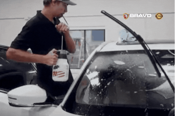 A professional installer at Bravo Protection sprays solution onto a white vehicle’s windshield during the early stage of applying windshield protection film, demonstrating the skill and precision behind expert service. The scene supports the importance of professional care outlined in Why Professional Installation Matters.