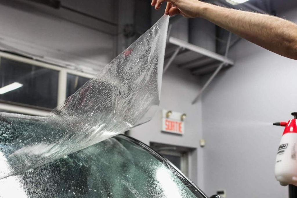 A person peels back a transparent windshield protection film from a car’s glass surface inside a Minnesota garage, demonstrating the flexible and durable qualities of high-performance TPU film. This visual supports the breakdown of different Types of Windshield Protection Film.